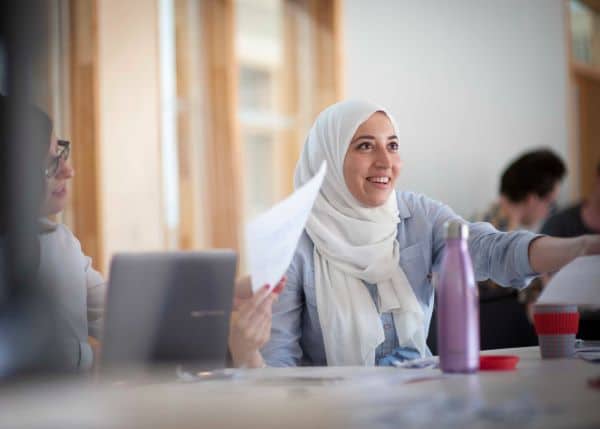 Image of a person holding papers, pointing off camera, they are wearing a white headscarf and there are desks in the foreground
