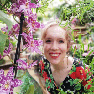 A white woman with light brown hair in a high bun, smiling while posing behind some flowers.