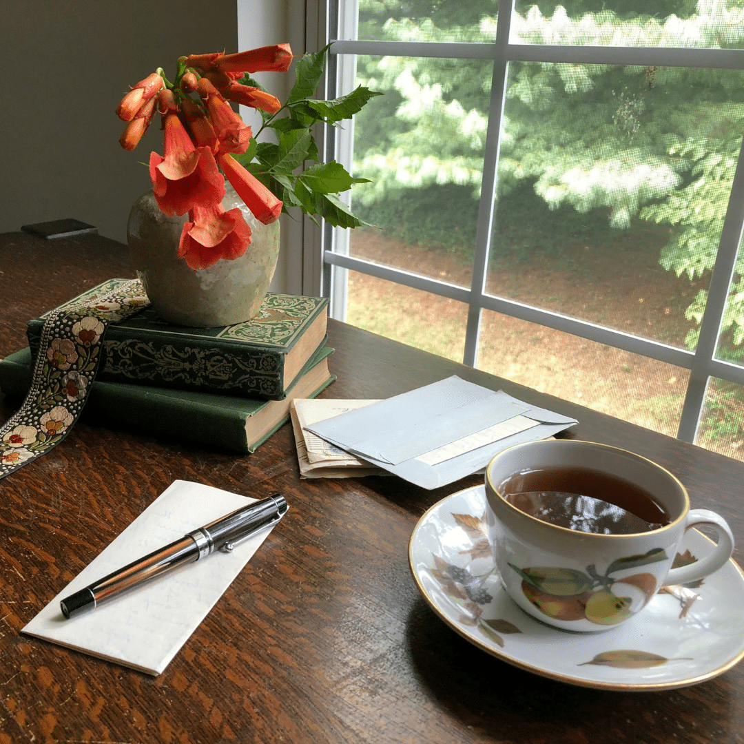 A cup of tea or coffee and a stack of letters await. In the background, a vase of orange trumpet vine flowers on a stack of books oversee the work as light streams through a paned window. Photo by Diane Helentjaris on Unsplash